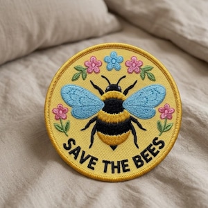 May include: Round yellow embroidered patch featuring a bee with blue wings, black and yellow body, and surrounded by pink and blue flowers. The text "SAVE THE BEES" is embroidered in black around the bee.