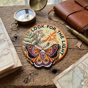 May include: Embroidered patch with a moth design in shades of orange, pink, and black, surrounded by green foliage and the text "LOOK FOR THE LIGHT" in a circular shape. The patch is on a wooden surface with a compass, journal, and map.
