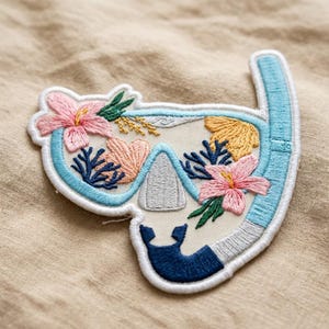 May include: Embroidered patch featuring a snorkeling mask design. The mask is outlined in light blue with a white border and filled with embroidered flowers in pink, coral, and yellow. Dark blue coral and green leaves add detail to the design.