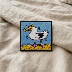 May include: Embroidered patch featuring a cartoon seagull with a yellow snack in its beak. The patch has a blue background and a yellow and black border. The seagull is white with black accents and pink legs.