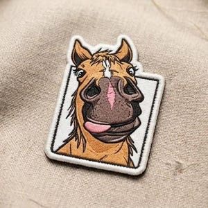 May include: Embroidered patch featuring a cartoon horse with a surprised expression. The horse has a brown mane and ears, a pink tongue, and a white rectangle border. The patch is on a beige fabric.