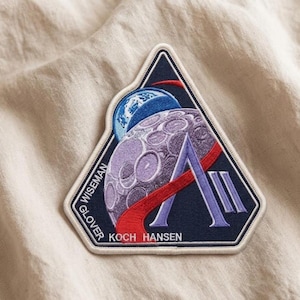 May include: Embroidered patch featuring a space-themed design with a purple moon, blue planet, and red accents. The patch is triangular with the names WISEMAN, GLOVER, KOCH, and HANSEN printed on it.
