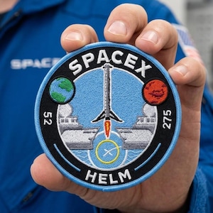 May include: A round SpaceX patch with a blue background, featuring a rocket launching upwards. The patch includes the word "SPACEX" in white, along with a green Earth and a red Mars. The word "HELM" is at the bottom.