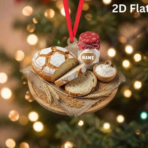 May include: A 2D flat hanging ornament featuring a round wooden board with a loaf of crusty bread, a slice of bread, a small bowl of flour, and a glass jar with a red patterned lid labeled NAME, all resting on a piece of burlap fabric.