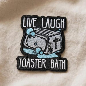 May include: Embroidered patch with the words "LIVE LAUGH" above a gray toaster in a blue bubbly bath, and the words "TOASTER BATH" below. The patch has a black border and is on a beige background.