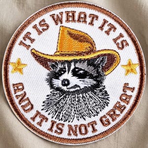 May include: Embroidered patch featuring a raccoon wearing a cowboy hat. The patch is circular with the text "IT IS WHAT IT IS AND IT IS NOT GREAT" in a brown border. Two gold stars are on either side of the raccoon.