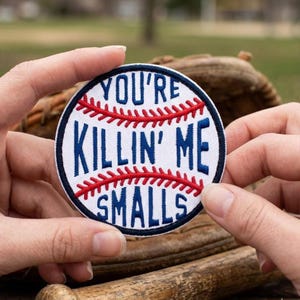 May include: A round patch with the text "YOU'RE KILLIN' ME SMALLS" in blue lettering. The patch has a baseball design with red stitching and a white background. The patch is held in front of a baseball glove.