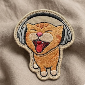 May include: Embroidered patch featuring a cartoon orange cat wearing headphones. The cat has a pink nose, open mouth, and a happy expression. The patch is outlined in a tan color, with a tan background.