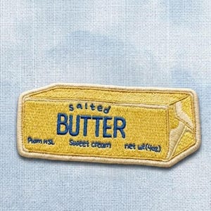 May include: A rectangular embroidered patch shaped like a stick of butter with a yellow background and blue stitched text that reads salted BUTTER, Sweet cream, and net wt (4oz). The patch has a light cream-colored border and textured stitching.
