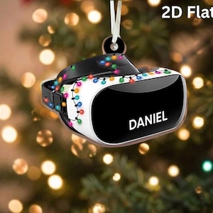 May include: A 2D flat ornament shaped like a virtual reality headset, featuring a black and white design wrapped in colorful Christmas string lights. The front of the headset displays the name DANIEL in white capital letters, with a white ribbon loop.