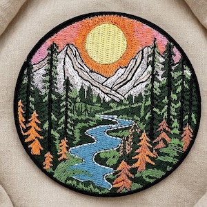 May include: Embroidered patch featuring a mountain landscape with a river, trees, and a setting sun. The design includes a blue river, green and orange trees, and a yellow sun against a pink and orange sky. The patch is circular with a black border.