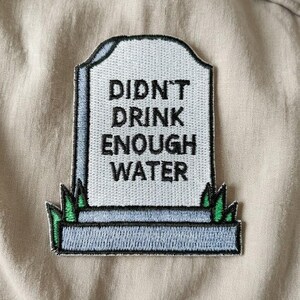 May include: Embroidered patch in the shape of a tombstone with the words "DIDN'T DRINK ENOUGH WATER" in black. The tombstone is light gray with a dark gray border and green grass at the bottom. The patch is on a tan fabric.