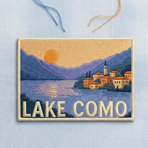 May include: A rectangular embroidered patch featuring a landscape of Lake Como with a golden sun, purple mountains, and a cluster of orange-roofed buildings by the water. The text LAKE COMO is stitched in large, bold, cream-colored capital letters.