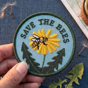 May include: Round embroidered patch with the text "SAVE THE BEES" in black lettering. The patch features a yellow flower with a bee and green leaves on a light blue background, with a green border. The patch is held by a hand.