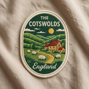 May include: An oval embroidered patch featuring a landscape of The Cotswolds, England. The design shows a stone house with a red roof, rolling green hills, a yellow sun, and four white sheep grazing near a winding path, all bordered by cream.