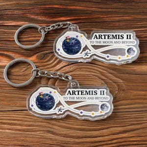 May include: Two clear acrylic keychains with silver key rings and chains. Each keychain features a design with a blue planet, a moon, and the text "ARTEMIS II TO THE MOON AND BEYOND". The keychains are on a wooden surface.