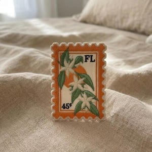 May include: Embroidered patch in the shape of a postage stamp. The patch features an orange border with a white and orange center. The center design includes white flowers, green leaves, and the letters "FL". The price is marked as 45¢.