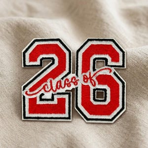 May include: A red and white embroidered patch with the number "26" in a collegiate style. The patch has a black border and the words "class of" in a cursive font. The patch is on a beige fabric.