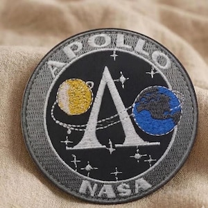 May include: Circular embroidered patch featuring the Apollo mission logo. The patch has a gray border, with the word "APOLLO" arched across the top. The NASA logo is at the bottom. The design includes a yellow planet, a blue planet, and the letter "A".