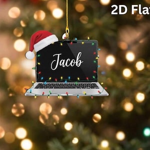 May include: A 2D flat hanging ornament shaped like a laptop with a silver base and black screen. The name Jacob is written in white script on the screen. A red and white Santa hat sits on the top left corner, and colorful Christmas lights wrap around the frame.