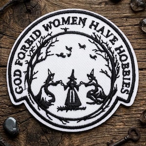 May include: A circular white patch with black embroidered text and imagery. The text reads "GOD FORBID WOMEN HAVE HOBBIES" around the border. The central image depicts three figures in a circle, with trees and birds.