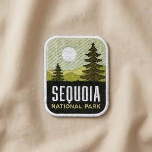 May include: Embroidered patch featuring a landscape with two pine trees, a sun, and the text "SEQUOIA NATIONAL PARK." The patch has a white border and is set against a tan fabric background.