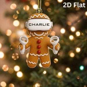May include: A flat gingerbread person ornament featuring white icing details, three red buttons, and a white virtual reality headset with the name CHARLIE printed in black text. The figure holds two white handheld controllers, one in each hand.