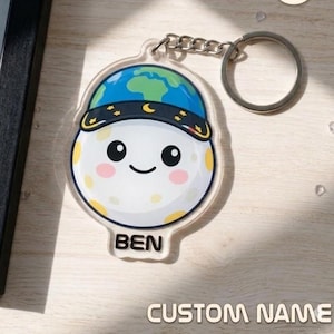 May include: A clear acrylic keychain featuring a smiling moon character wearing a blue and green earth-themed hat. The keychain has a silver keyring and chain. The name "BEN" is printed on the keychain. The words "CUSTOM NAME" are printed below.