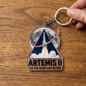 May include: A clear acrylic keychain featuring the Artemis II mission. The design includes a rocket launching against a starry sky and a full moon. The keychain has a silver chain and ring. The text "ARTEMIS II TO THE MOON AND BEYOND" is printed on the keychain.