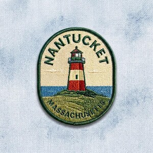 May include: An oval embroidered patch featuring a lighthouse with red and white stripes on a grassy hill. The text NANTUCKET is arched above the lighthouse, and MASSACHUSETTS is curved below. The design includes a blue sea and a cream background.