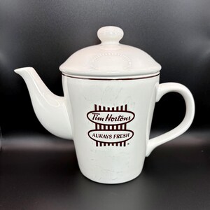 May include: White ceramic teapot with a lid and handle. The teapot features the Tim Hortons logo in brown with the words "ALWAYS FRESH". The teapot has a spout and a handle.