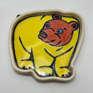May include: A hand-painted ceramic dish shaped like a bear. The bear has a yellow body, a red head, and blue eyes. The dish has a cream-coloured rim and black outlines. The bear is depicted in a playful pose.