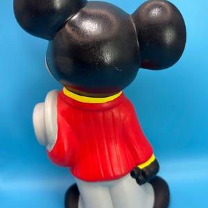 Peut inclure: Figurine en c&eacute;ramique d'un personnage de dessin anim&eacute; de souris, vue de dos. La figurine a de grandes oreilles noires, une veste rouge avec une bordure jaune, un pantalon gris et des chaussures noires. Un chapeau blanc est tenu sur le c&ocirc;t&eacute; gauche. Sur fond bleu.