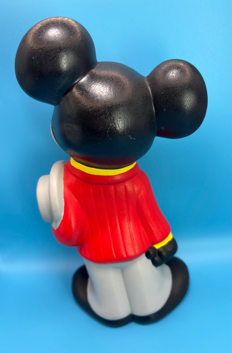 Peut inclure: Figurine en c&eacute;ramique d'un personnage de dessin anim&eacute; de souris, vue de dos. La figurine a de grandes oreilles noires, une veste rouge avec une bordure jaune, un pantalon gris et des chaussures noires. Un chapeau blanc est tenu sur le c&ocirc;t&eacute; gauche. Sur fond bleu.