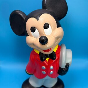 Peut inclure: Figurine en c&eacute;ramique de Mickey Mouse, v&ecirc;tu d'une veste rouge, d'un pantalon gris et de chaussures noires. Il porte un n&oelig;ud papillon noir, un col jaune et tient un chapeau gris. Le fond est bleu uni.
