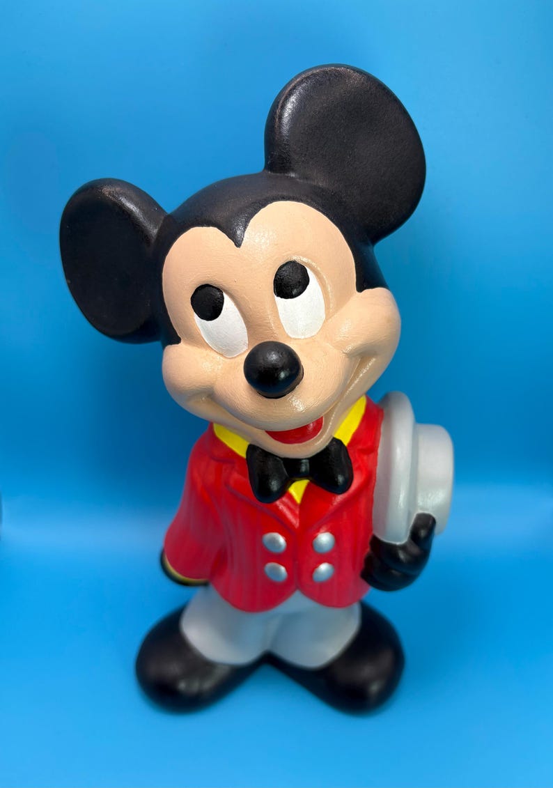 Peut inclure: Figurine en c&eacute;ramique de Mickey Mouse, v&ecirc;tu d'une veste rouge, d'un pantalon gris et de chaussures noires. Il porte un n&oelig;ud papillon noir, un col jaune et tient un chapeau gris. Le fond est bleu uni.