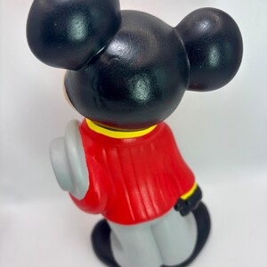 Peut inclure: Figurine vintage de Mickey Mouse, vue de dos, avec de grandes oreilles noires et une veste rouge. La figurine tient un chapeau gris et porte un pantalon gris. La veste a des bordures jaunes et noires. La figurine est sur fond blanc.