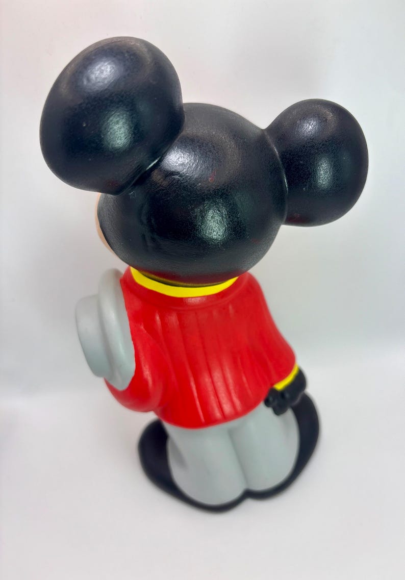 Peut inclure: Figurine vintage de Mickey Mouse, vue de dos, avec de grandes oreilles noires et une veste rouge. La figurine tient un chapeau gris et porte un pantalon gris. La veste a des bordures jaunes et noires. La figurine est sur fond blanc.