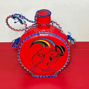 May include: Red leather flask with a cartoon squirrel graphic. The flask has blue and white stitching and a braided strap. The squirrel graphic features a rainbow and is outlined in black. The flask is a novelty item.
