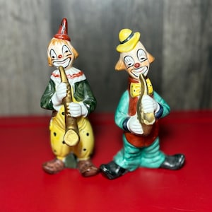 Peut inclure: Deux figurines de clown en céramique. L'un porte une tenue rouge et verte avec un pantalon jaune à pois, l'autre porte une tenue verte et jaune. Les deux clowns tiennent un instrument de musique doré et ont des visages peints avec des sourires.