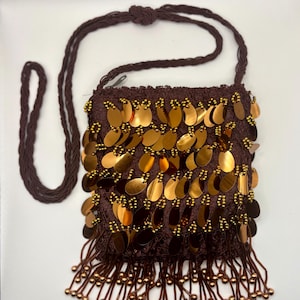 May include: A brown shoulder bag with gold sequins and beaded fringe. The bag has a woven texture and a long, braided strap. The sequins are oval-shaped and arranged across the front of the bag.