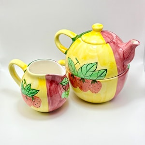 Può includere: Un set di teiera e lattiera in ceramica. La teiera e la lattiera sono dipinte con una combinazione di colori giallo, rosso e verde. La teiera e la lattiera sono decorate con disegni di fragole e foglie.