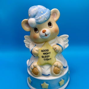 Puede incluir: Figura de cerámica de un osito de peluche con gorro de dormir azul y sosteniendo una estrella con la frase "GOOD NIGHT SLEEP TIGHT". El oso tiene pequeñas alas de ángel y está sentado sobre una base azul con estrellas.