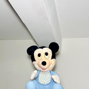 May include: A plush Mickey Mouse doll in a blue outfit and bib with a white polka dot pattern. The doll has black ears and a cream-coloured face. It is sitting on a red marquee letter.