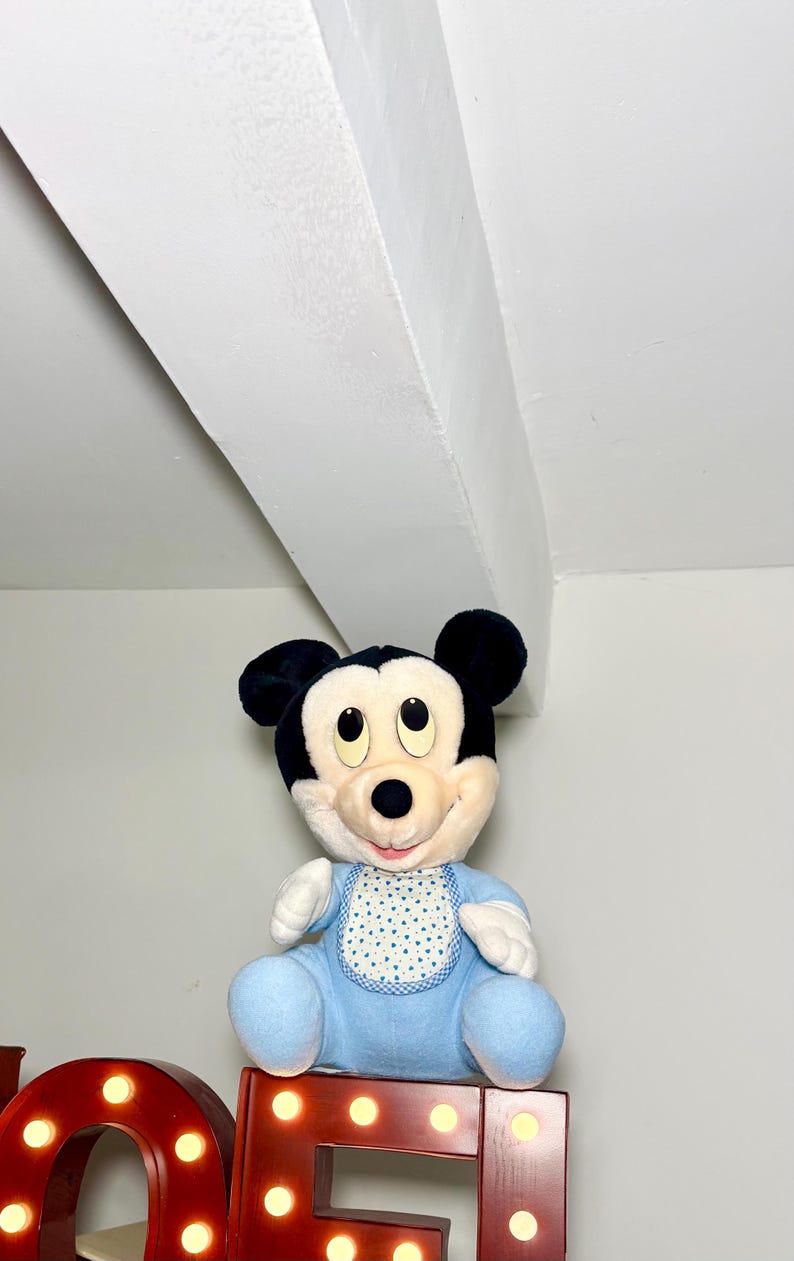 May include: A plush Mickey Mouse doll in a blue outfit and bib with a white polka dot pattern. The doll has black ears and a cream-coloured face. It is sitting on a red marquee letter.
