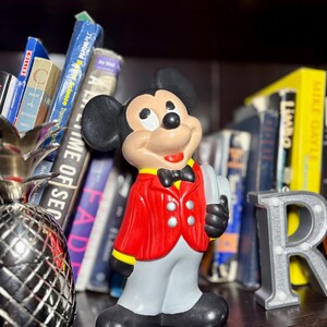 Peut inclure: Figurine en c&eacute;ramique de Mickey Mouse portant une veste rouge, un pantalon gris et des chaussures noires. La figurine tient un plateau et porte un n&oelig;ud papillon noir. Des livres et une lettre "R" argent&eacute;e sont en arri&egrave;re-plan.