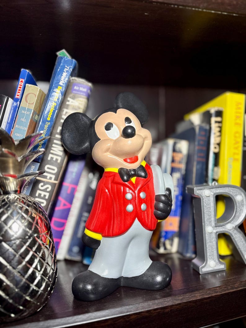 Peut inclure: Figurine en c&eacute;ramique de Mickey Mouse portant une veste rouge, un pantalon gris et des chaussures noires. La figurine tient un plateau et porte un n&oelig;ud papillon noir. Des livres et une lettre "R" argent&eacute;e sont en arri&egrave;re-plan.