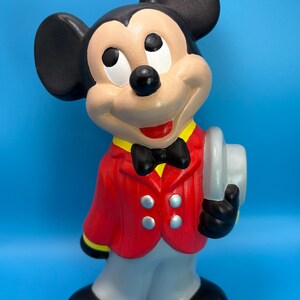 Peut inclure: Figurine en c&eacute;ramique de Mickey Mouse, v&ecirc;tu d'une veste rouge, d'un pantalon gris et de chaussures noires. La figurine porte un n&oelig;ud papillon noir, des oreilles noires et tient un chapeau gris. Le fond est bleu uni.