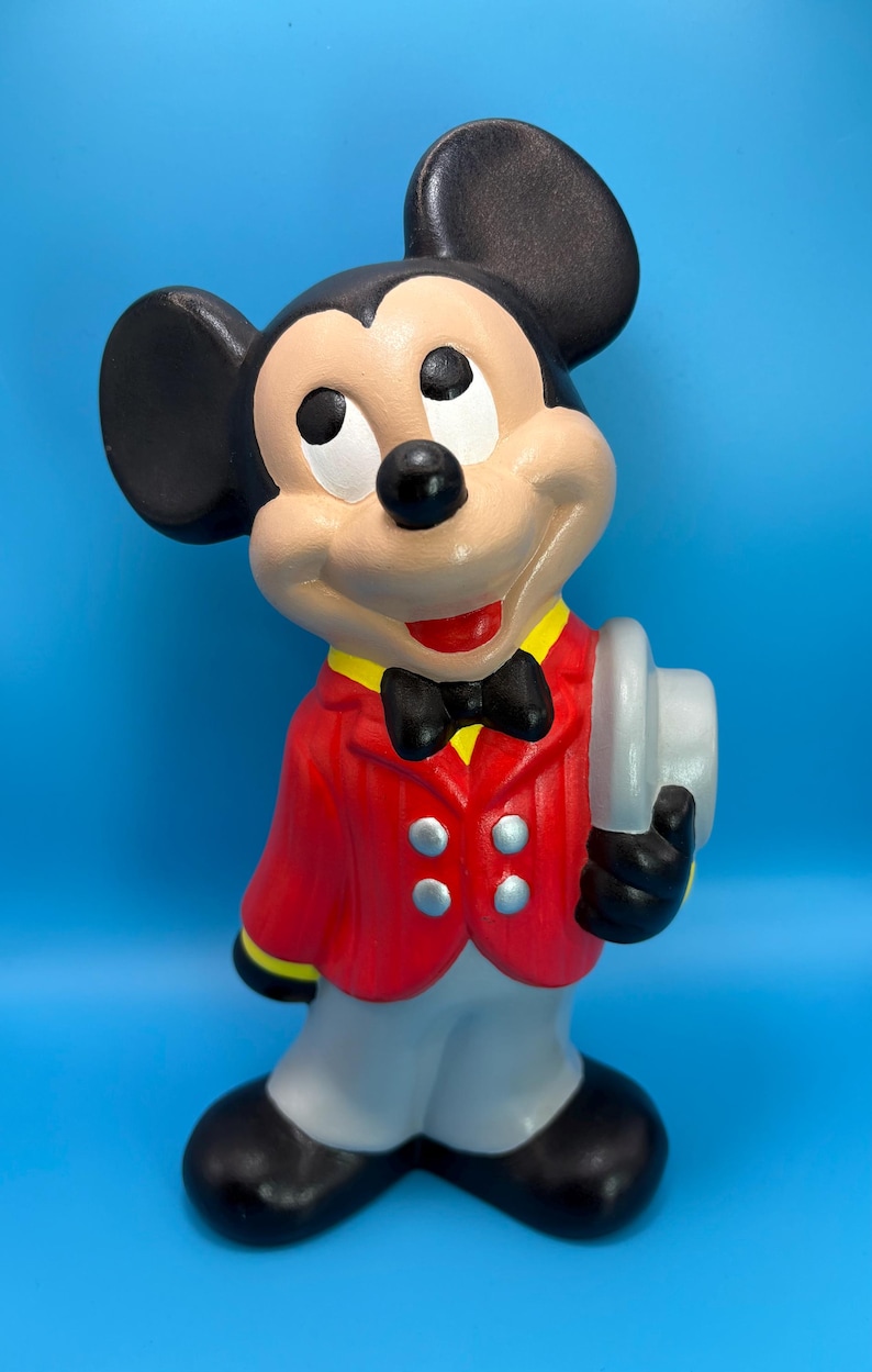 Peut inclure: Figurine en c&eacute;ramique de Mickey Mouse, v&ecirc;tu d'une veste rouge, d'un pantalon gris et de chaussures noires. La figurine porte un n&oelig;ud papillon noir, des oreilles noires et tient un chapeau gris. Le fond est bleu uni.