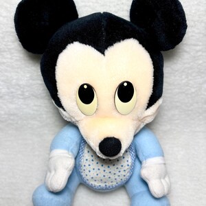 May include: A soft toy Mickey Mouse with black ears and head. The doll has a white face and a blue outfit with a bib featuring a white heart pattern. The doll's eyes are large and black.